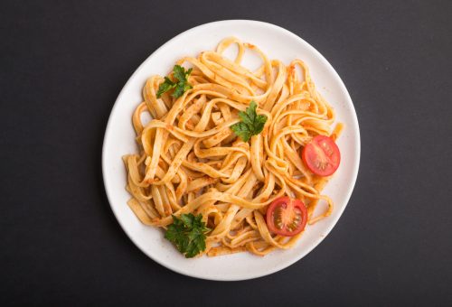Semolina pasta with tomato pesto sauce, orange and herbs on a white ceramic plate on a black background. Top view, flat lay, close up.